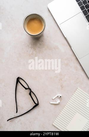 Minimal workspace with laptop, black glasses, coffee cup, wireless earbuds, and striped notebook on white desk - modern professional setup. Top view. Stock Photo