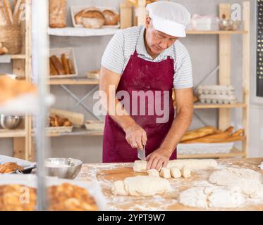 Senior man works in bakery as baker, cuts dough into portions, forms pieces of dough to create buns. Stock Photo