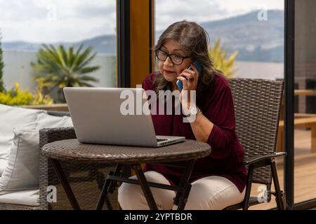 Elderly woman talking on cell phone while looking at her laptop on a terrace in the morning Stock Photo