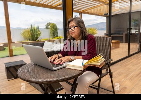 Senior Latina woman with glasses working with a laptop and a notebook on a terrace with vegetation Stock Photo