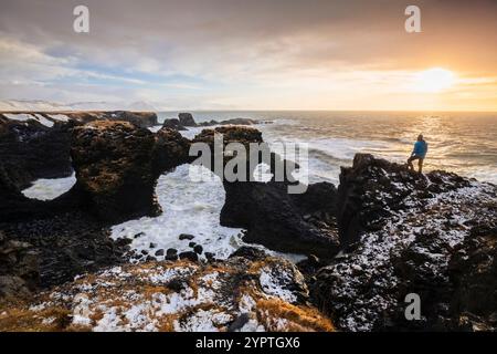 View of a man admiring the Gatklettur rock arch at sunrise in winter. Arnarstapi, Hellnar, Snæfellsnes Peninsula, Iceland, Northern Europe. Stock Photo