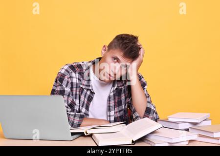 Young student with books and laptop having stress before exam at table against yellow background Stock Photo