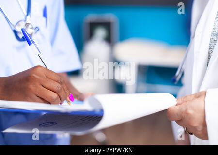 Closeup on hands of caucasian doctor and black nurse holding clipboard in medical office. Selective focus on african american person using pen to write medical diagnosis and prescriptions for patient. Stock Photo
