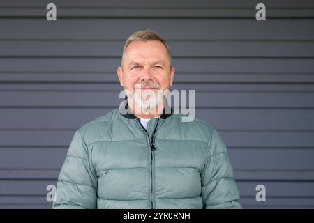 A middle-aged man with a gray beard and a gentle smile stands confidently against a plain background, wearing a light green puffer jacket. Stock Photo