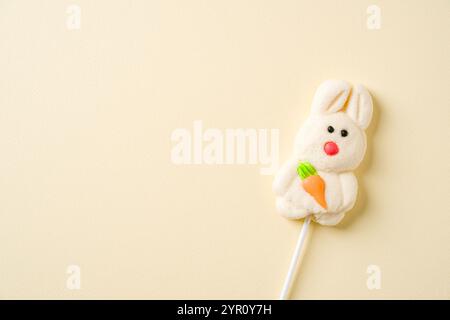 A cute bunny shaped marshmallow lollipop holding an orange carrot, set on a minimalist pastel background Stock Photo