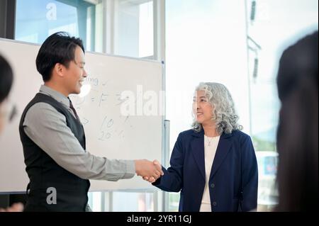 A successful senior Asian female CEO shaking hands with a young, smart male CEO during a collaborative business meeting in front of their employees in Stock Photo