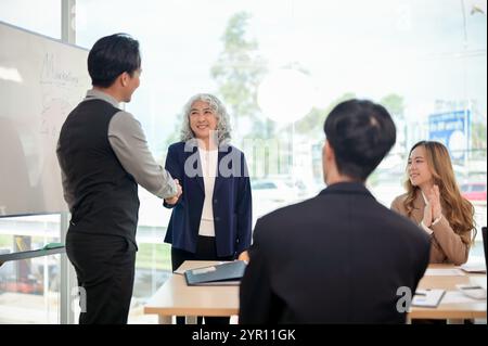A successful senior Asian female CEO shaking hands with a young, smart male CEO during a collaborative business meeting in front of their employees in Stock Photo