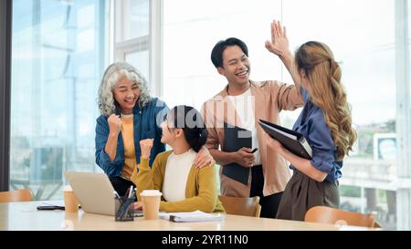 A group of smiling, cheerful, diverse Asian businesspeople celebrates their project success in the office, raising their fists in excitement and givin Stock Photo