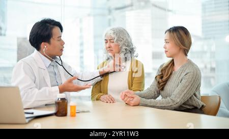 A retired, concerned Asian grandmother having her internal body sounds checked with a stethoscope while visiting the doctor with her granddaughter at Stock Photo