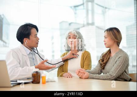 A retired, concerned Asian grandmother having her internal body sounds checked with a stethoscope while visiting the doctor with her granddaughter at Stock Photo