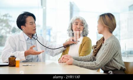A retired, concerned Asian grandmother having her internal body sounds checked with a stethoscope while visiting the doctor with her granddaughter at Stock Photo
