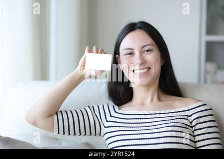 Happy asian woman showing blank card sitting on a couch at home Stock Photo