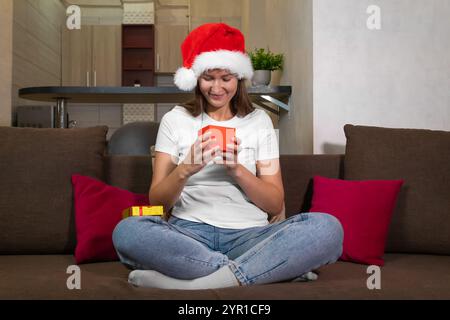 Young woman in a Santa Claus hat sitting on the couch opens a small red Christmas gift box and smiles, anticipating the surprise inside Stock Photo