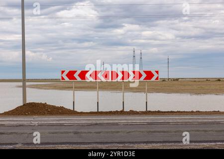 Red arrow signs warn drivers of a sharp curve ahead on a road adjacent to a flooded area under a cloudy sky Stock Photo