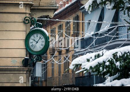 A vintage green clock with Roman numerals is mounted on the corner of a building in a snowy, historic European city street in Alba, Italy. Stock Photo