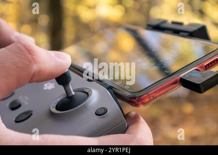 Kharkiv, Ukraine - October 15, 2021: Dji drone remote controller in hands with connected phone. Operating drone close-up in autumn golden forest blurr Stock Photo