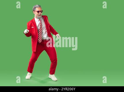 Happy dancing grey haired man in red suit and sunglasses in Santa style on green background. Stock Photo