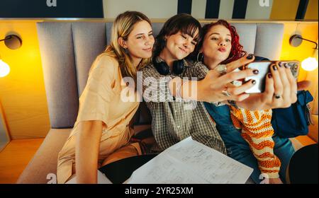 A group of female students enjoying a playful moment while taking a selfie in a warm, inviting study setting, surrounded by notes and cozy lighting. Stock Photo