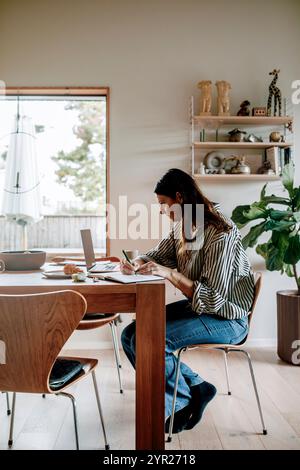 Focused female freelancer working while sitting near dining table in living room at home Stock Photo