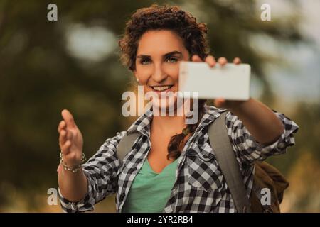 Young female hiker with a backpack takes a joyful selfie in nature, embodying the spirit of exploration and sharing moments on social media Stock Photo
