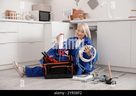 Mature female plumber sitting on floor with bag of tools in kitchen Stock Photo