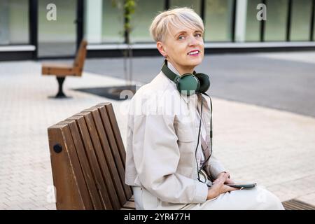 Beautiful Smiling Mature Woman with Short Hair Wearing Headphones and Using phone While Sitting on Bench Outdoors in City, Active lifestyle for Older Stock Photo