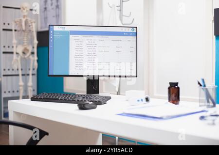 Empty doctors office with healthcare equipment and cabinet neatly arranged for use. Computer displaying medical data is on a desk in clinical room, along with a clipboard, thermometer and pill bottle. Stock Photo
