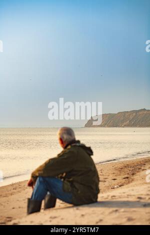 A man from behind, sitting on the beach and looking out to sea Stock Photo