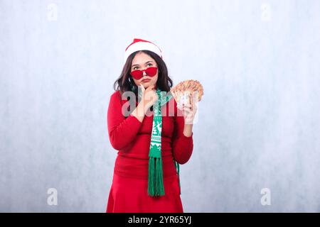 gesture of young Asian ladies wearing red christmas sweater, red glasses, Santa hat and scarf, seriously thinking hand holding euro cash money and chi Stock Photo