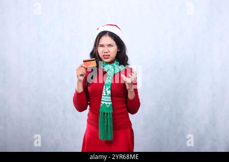 gesture of young asian ladies wearing red christmas sweater, santa hat and scarf, upset to camera hand holding credit debit card and growling emotion, Stock Photo