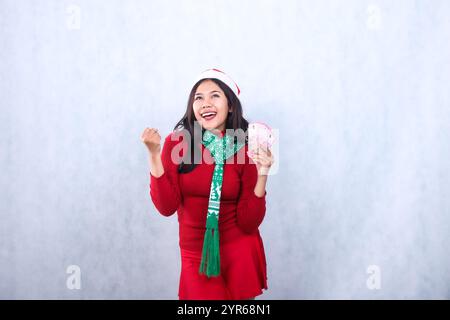 gesture of young Asian ladies wearing red christmas sweater, Santa hat, and scarf, cheerful up hands holding rupiah cash money and clenched successful Stock Photo