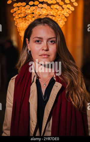 Vertical. Serious young woman with long blonde hair and a red scarf, standing at night under festive lights, looking at camera with a focused expression, Lady with pensive attitude staring front. High quality photo Stock Photo