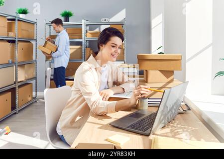 Female manager working on laptop, man doing inventory in warehouse, inspecting boxes and parcels. Stock Photo