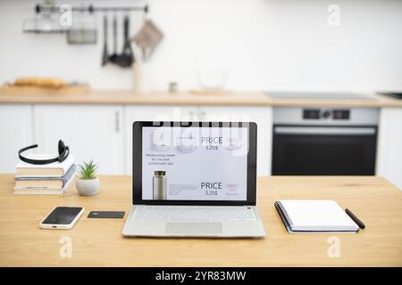 Laptop on desk shows online cosmetics store offering skincare products. Modern kitchen setting features wooden table with notebook, smartphone, and pen. Stock Photo
