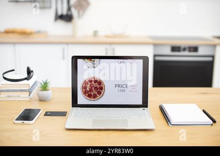 Modern kitchen setting with laptop displaying fast food online sales site featuring pizza. Smartphone, credit card, and notebook on wooden table. Stock Photo