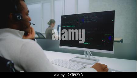 African American professional stockbroker in headset analyzes financial charts and diagrams on computer. PC screen shows stock market and shares. Modern office of stock exchange company. E-commerce. Stock Photo