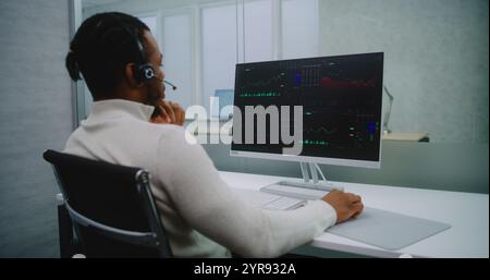 African American professional stockbroker in headset analyzes financial charts and diagrams on computer. PC screen shows stock market and shares. Modern office of stock exchange company. E-commerce. Stock Photo