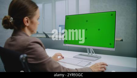 Professional female financial expert or marketing specialist enters data using computer with green screen chroma key. Mock up template for screen content on PC. Modern office of E-business company. Stock Photo