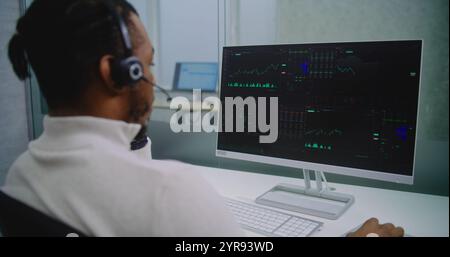 African American professional financial specialist in headset analyzes share market and stocks on computer. PC screen shows charts and diagrams. Modern office of stock exchange company. E-business. Stock Photo