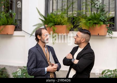 Two businessmen having a conversation outside a building, one holding a laptop, suggesting a work related discussion Stock Photo