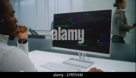 African American man works in modern office of international company: Stock exchange trader wearing headset monitors equity market and shares using computer, analyzes charts and diagrams. E-business. Stock Photo