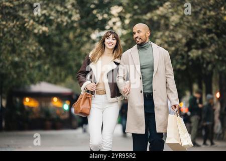 A joyful couple spending quality time shopping together, walking arm in arm down a tree-lined city street. Stock Photo