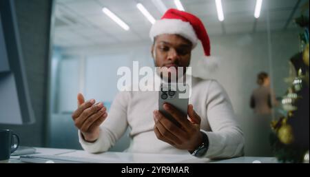 Young African American businessman in Santa hat talks to business partner about deal on video chat on mobile phone. Marketing specialist works in modern office decorated for Christmas. E-commerce. Stock Photo