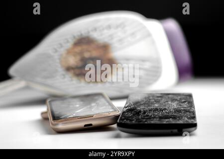 Old damaged smartphones with shattered cracked touch screens on a repair shop white table. Burned household steam electric iron blurs in the backgroun Stock Photo