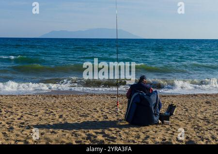 Man of retirement age sitting sea fishing on a sandy beach with ocean and island in the background Stock Photo