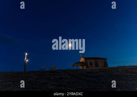 Small hilltop chapel illuminated under a full moon with a glowing statue in the deep blue night sky. Stock Photo