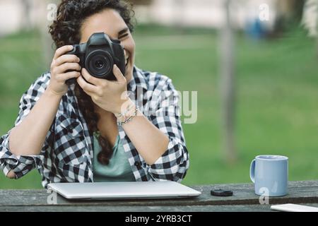 Young adult female photographer smiling while capturing images with a dslr camera in a park, laptop and coffee on the table, embracing her passion for Stock Photo
