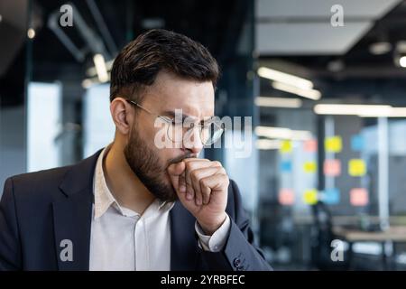 A businessman is seen coughing, experiencing cold or flu symptoms in an office environment. Wearing glasses and a suit, he holds his hand to his mouth, indicating illness and discomfort. Stock Photo