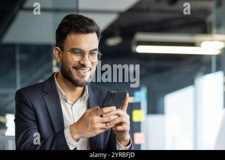 A man businessman is seated in an office environment, focused on his smartphone. His smile indicates happiness and connection, portraying a professional engaged in digital communication. Stock Photo