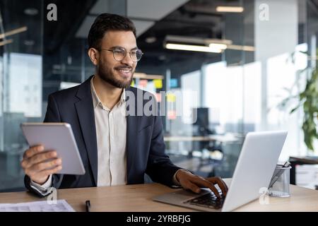 A businessman is seated in a modern office, using a laptop and tablet, embodying productivity and multitasking with digital devices. He displays confidence and a friendly demeanor Stock Photo